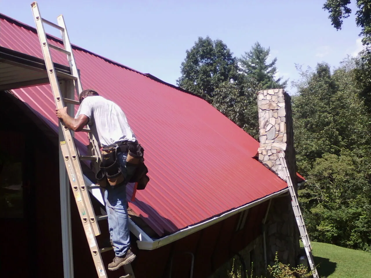 Roofer on a ladder beside a red metal roof and stone chimney — Soffit & Fascia Repair by Roofing Brothers in Corona